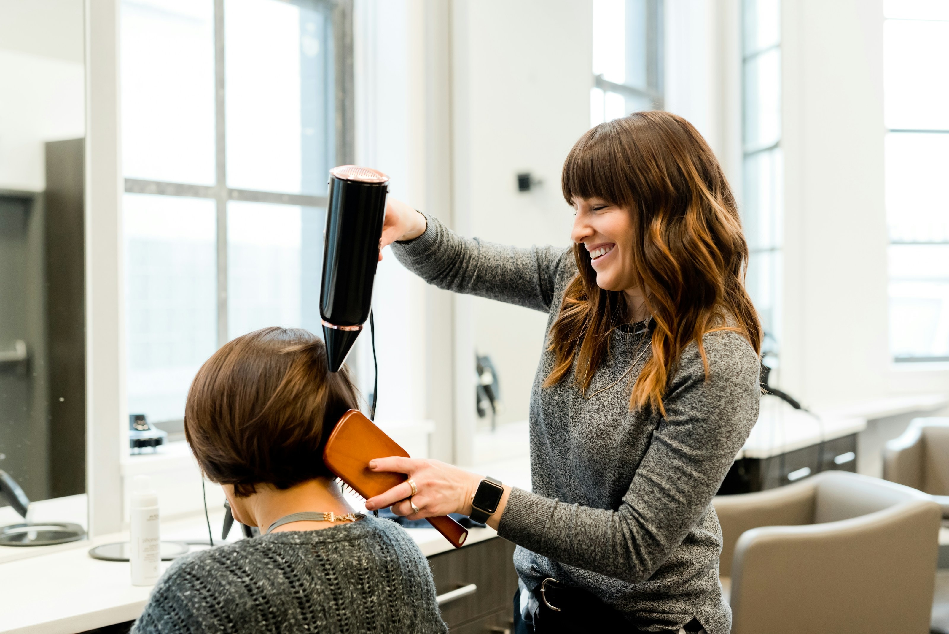 A smiling stylist blow-drying a customer's hair in a bright salon